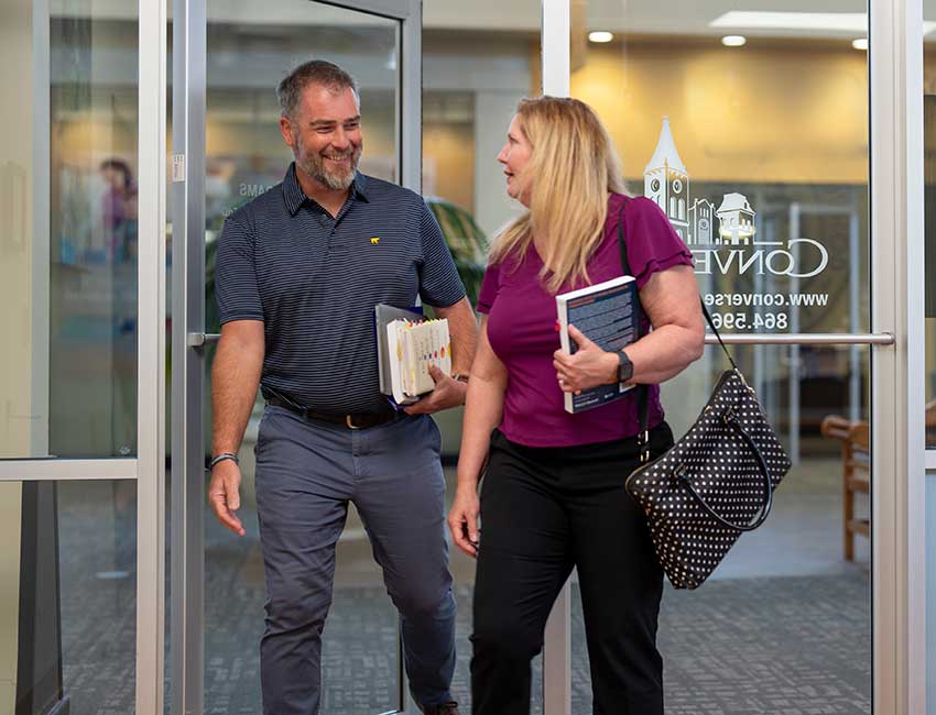 Two adult Converse Graduate students walk smiling into a Converse campus building .
