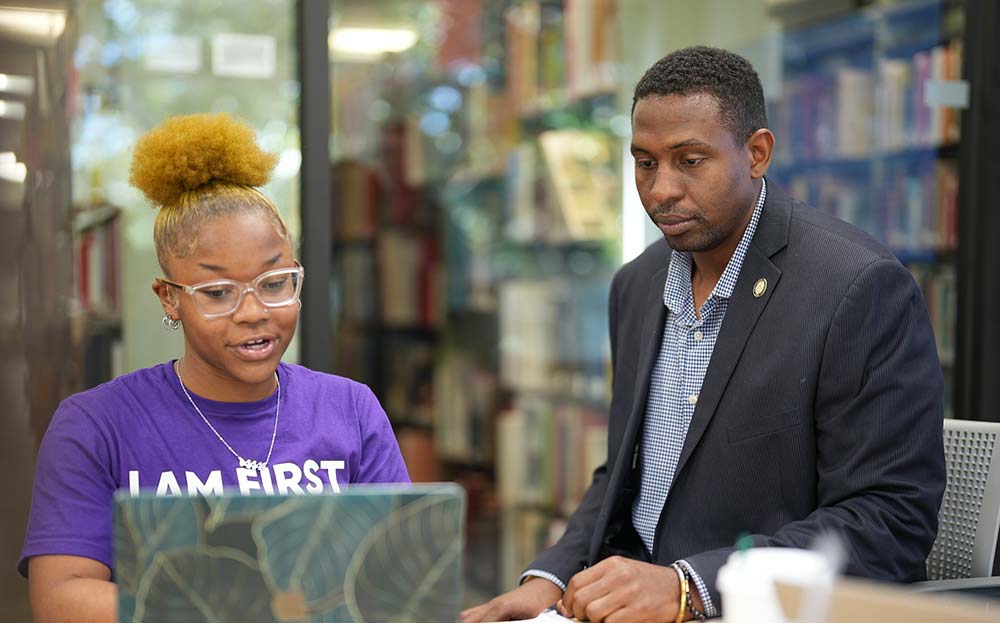 A faculty member guides a student as they work on their laptop.