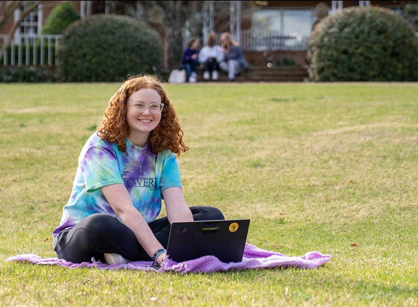 A Converse students works on a laptop on the campus quad on a sunny day.