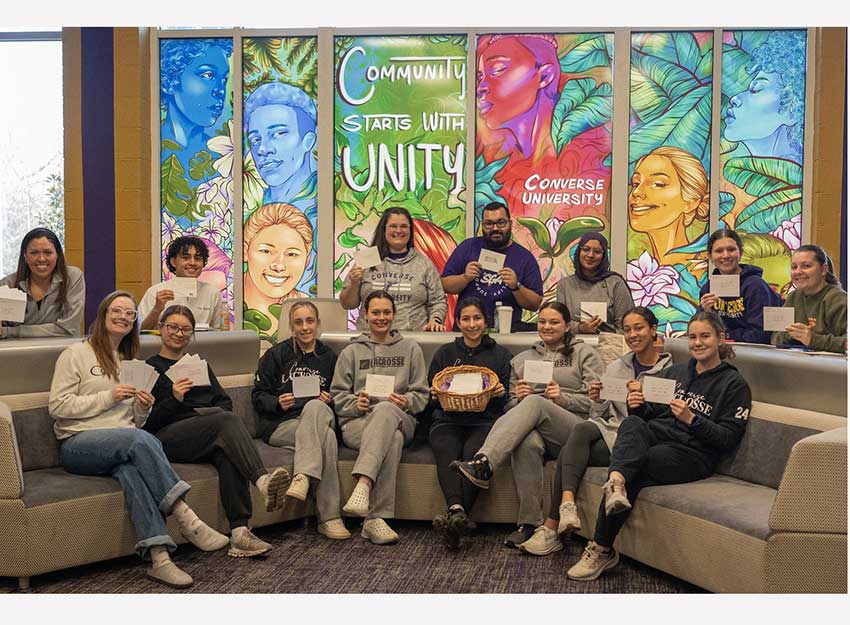 A group of volunteers sit in front of a unity mural holding up cards they wrote encouraging veterans and the eldery.