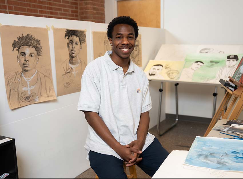 Joel Houston smiles sitting in an art studio in front of some of their work.