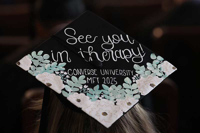 An MMFT student wearing a mortar board that says, "See you in therapy; Converse Univsitery MFT 2025
