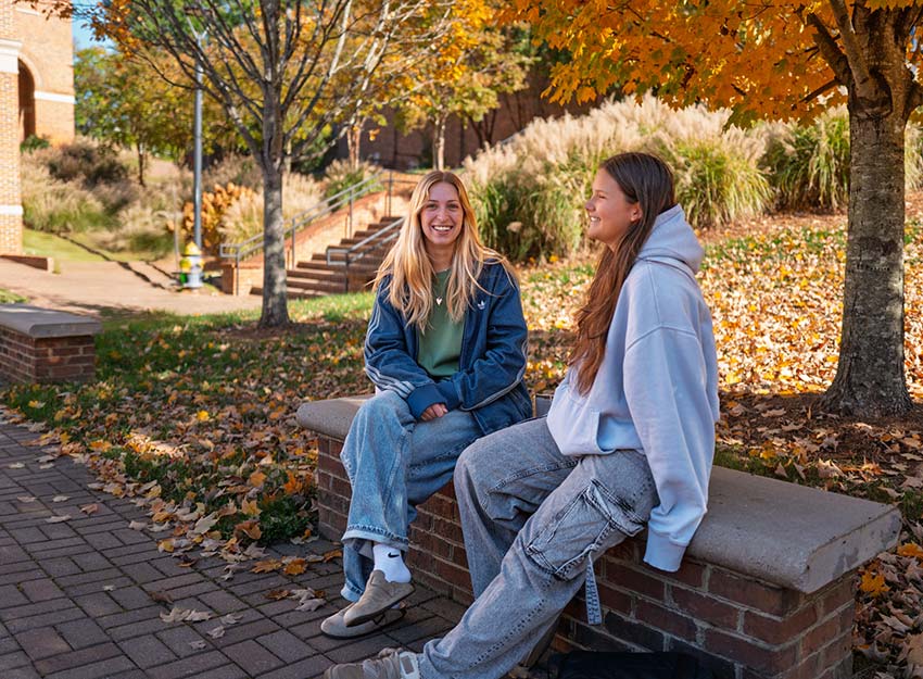 Two students sit outside on a fall day on campus talking and laughing.