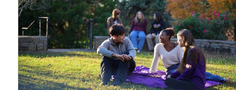 A group of Converse students sit on campus on a sunny day talking to each other.