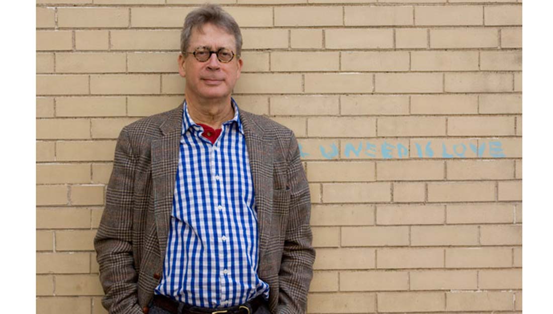 Tillinghast stands against a wall with writing that reads "all you need is love"