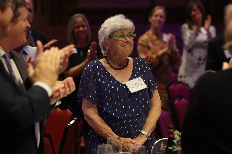 Gillian smiles proudly while people applaud around her.